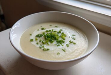 A white bowl filled with smooth, creamy potato leek soup, garnished with a swirl of cream and fresh chopped chives.