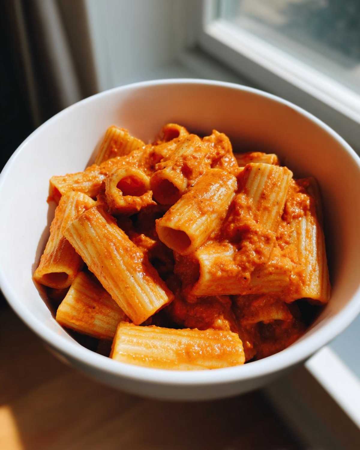 Close-up of rigatoni pasta coated in vibrant orange Creamy Roasted Red Pepper Pasta sauce in a white bowl.