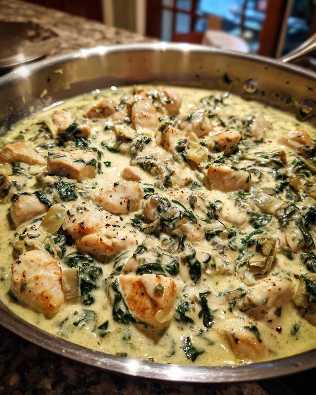 Close-up of chunks of seared chicken breast simmering in a rich, creamy spinach artichoke sauce in a stainless steel skillet.