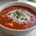 A close-up of a white bowl filled with rich, red Creamy Tomato Tortellini Soup, topped with shredded Parmesan cheese and fresh parsley.