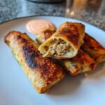 Four golden brown Crispy Baked Reuben Egg Rolls on a white plate, one cut open to show the filling, with a side of Thousand Island dip.