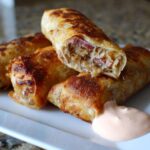 Close-up of crispy baked Reuben egg rolls, one cut open showing corned beef and sauerkraut, served with Thousand Island dip.