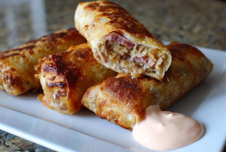 Close-up of crispy baked Reuben egg rolls, one cut open showing corned beef and sauerkraut, served with Thousand Island dip.