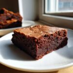 A close-up of a rich, fudgy Dark Chocolate Tahini Brownies slice with a crackly top, served on a white plate.