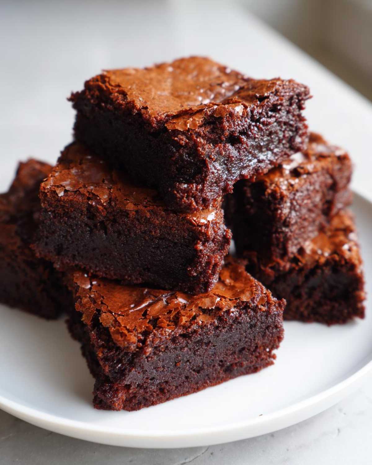 A stack of rich, fudgy Dark Chocolate Tahini Brownies with shiny, crackly tops served on a white plate.