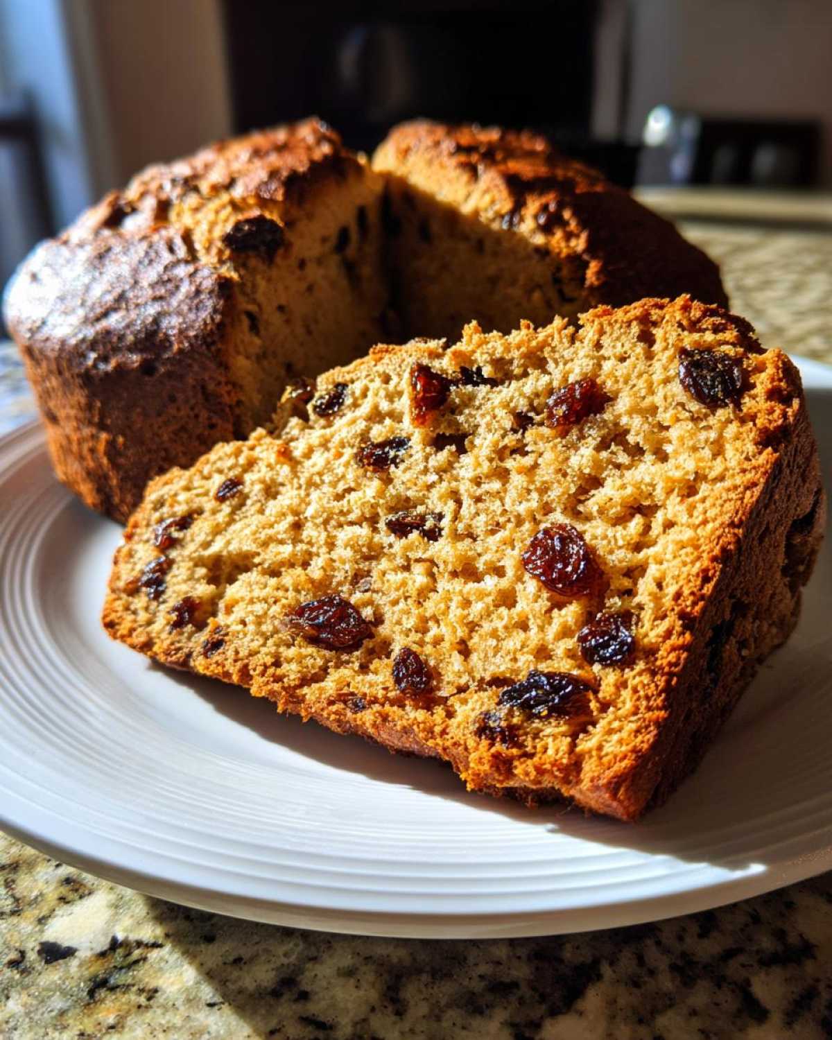 A thick slice of Easy Irish Soda Bread with Buttermilk and Raisins, showing a golden crumb and dark raisins, resting on a white plate.