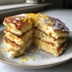 A close-up of a stack of fluffy Gluten-Free Buttermilk Pancakes with a bite taken out, topped with powdered sugar and orange zest.