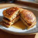 A stack of fluffy Vegan Banana Oat Pancakes cut in half, drizzled with maple syrup on a white plate.