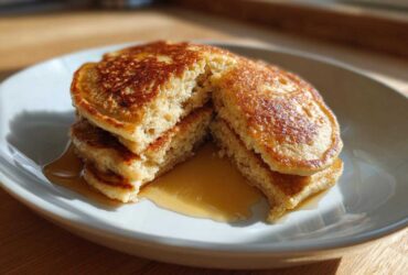 A stack of fluffy Vegan Banana Oat Pancakes cut in half, drizzled with maple syrup on a white plate.