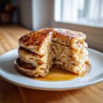 A tall stack of fluffy Vegan Banana Oat Pancakes with a slice cut out, drizzled generously with maple syrup.