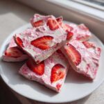 Close-up of several squares of Frozen Strawberry Yogurt Bark topped with fresh strawberry slices on a white plate.
