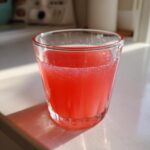 A glass filled with bright red liquid, likely Homemade Strawberry Lemonade Concentrate, sitting on a white counter in sunlight.