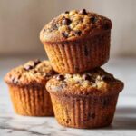 Three freshly baked Gluten-Free Banana Chocolate Chip Muffins stacked on a marble surface.