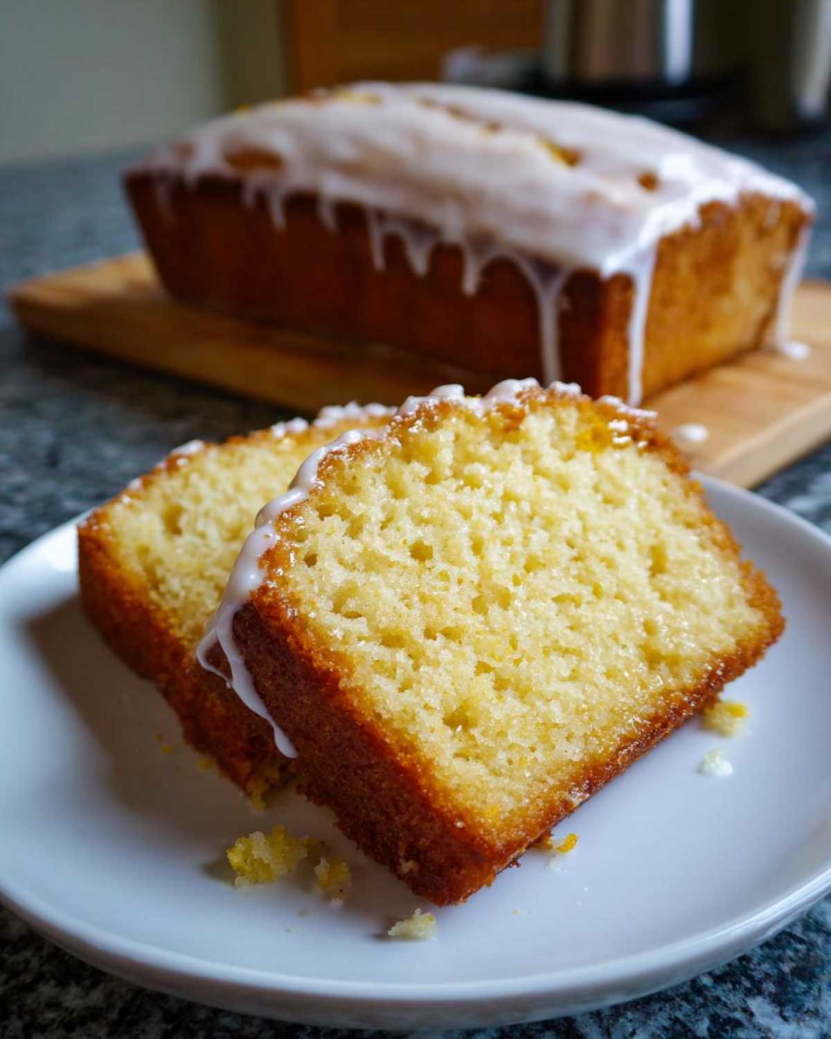 Two slices of moist Gluten-Free Lemon Coconut Cake topped with white lemon glaze, with the loaf in the background.
