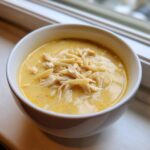 A close-up of a white bowl filled with rich, yellow Greek Lemon Chicken Soup featuring shredded chicken and orzo pasta.