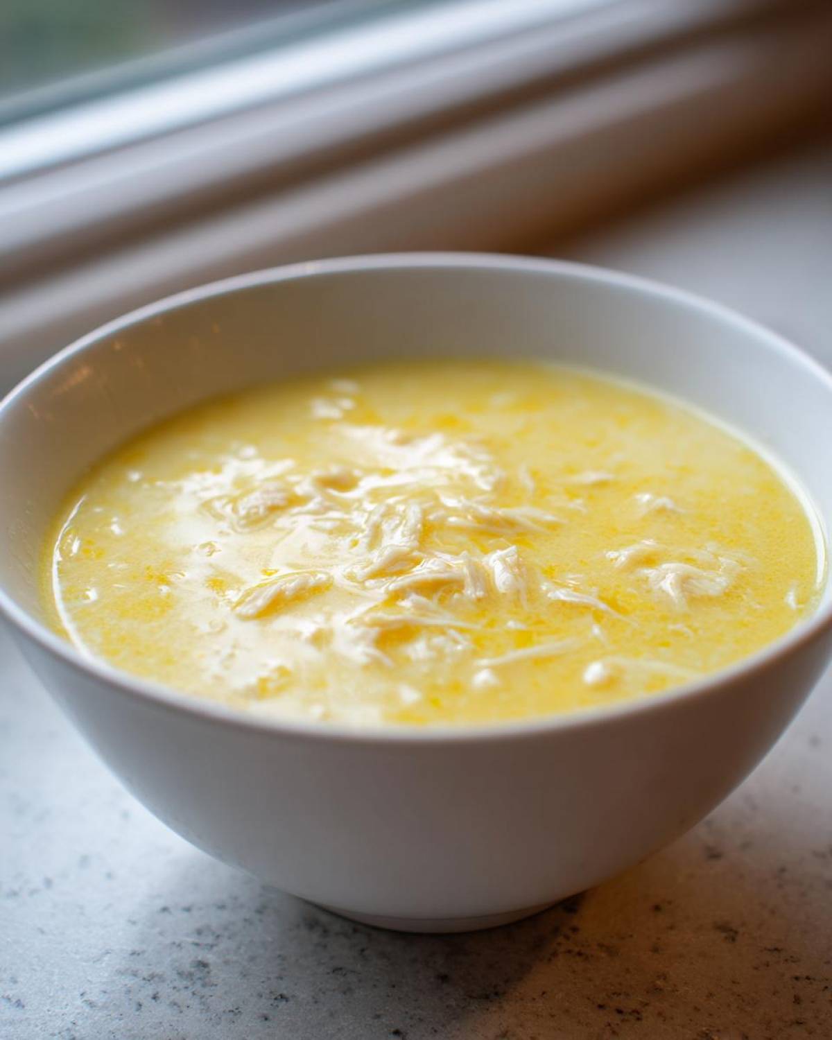 Close-up of a white bowl filled with creamy, yellow Greek Lemon Chicken Soup featuring shredded chicken.