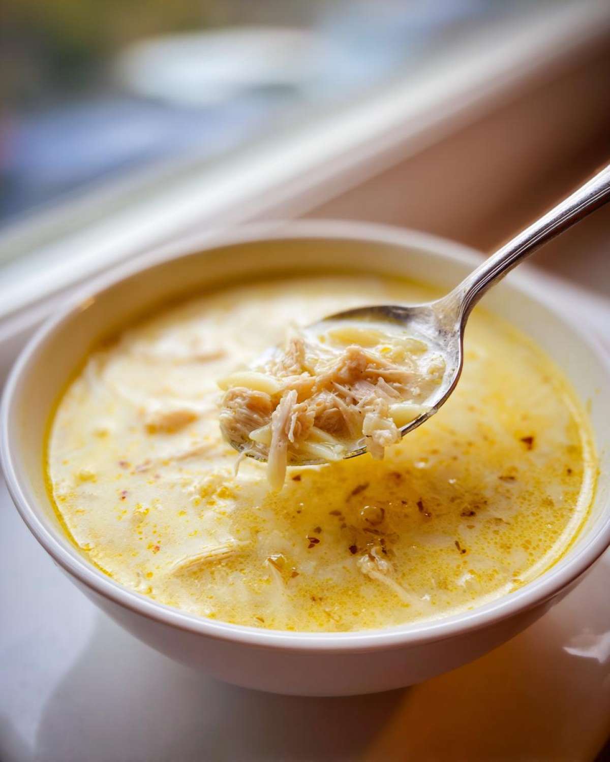 A spoonful of shredded chicken and noodles being lifted from a bowl of creamy Greek Lemon Chicken Soup.