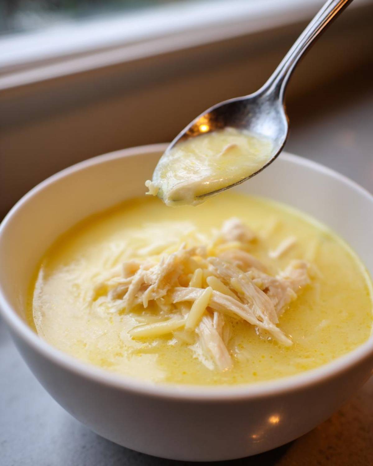 A spoonful of creamy Greek Lemon Chicken Soup being lifted from a white bowl topped with shredded chicken and orzo pasta.