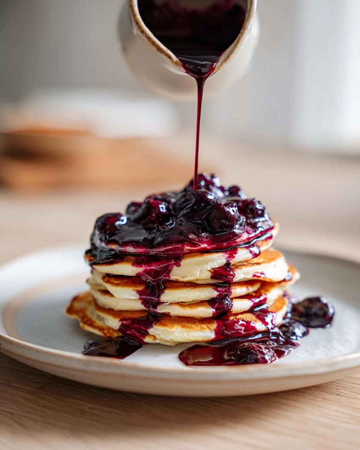 A stream of rich Homemade Blueberry Syrup is being poured from a small pitcher onto a stack of fluffy pancakes.