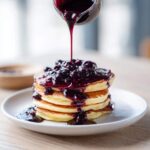 A stack of fluffy pancakes being generously topped with rich Homemade Blueberry Syrup.