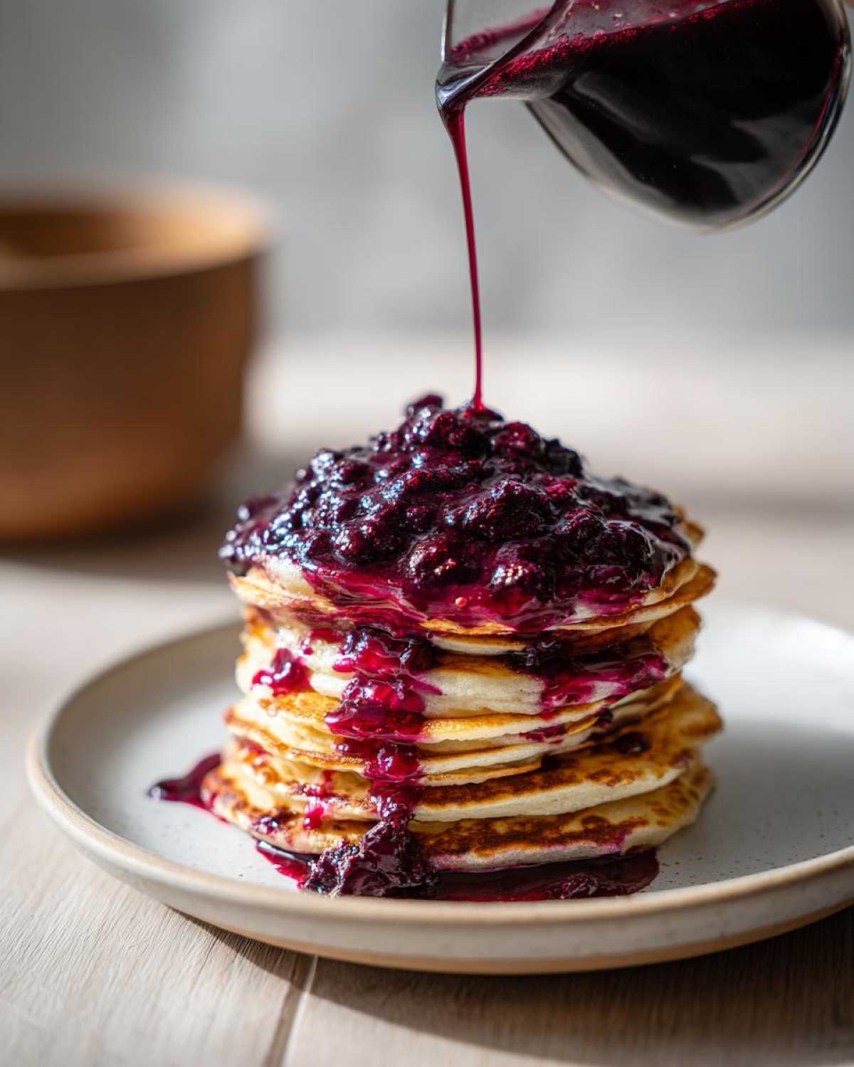 A stream of rich Homemade Blueberry Syrup being poured over a tall stack of golden pancakes.