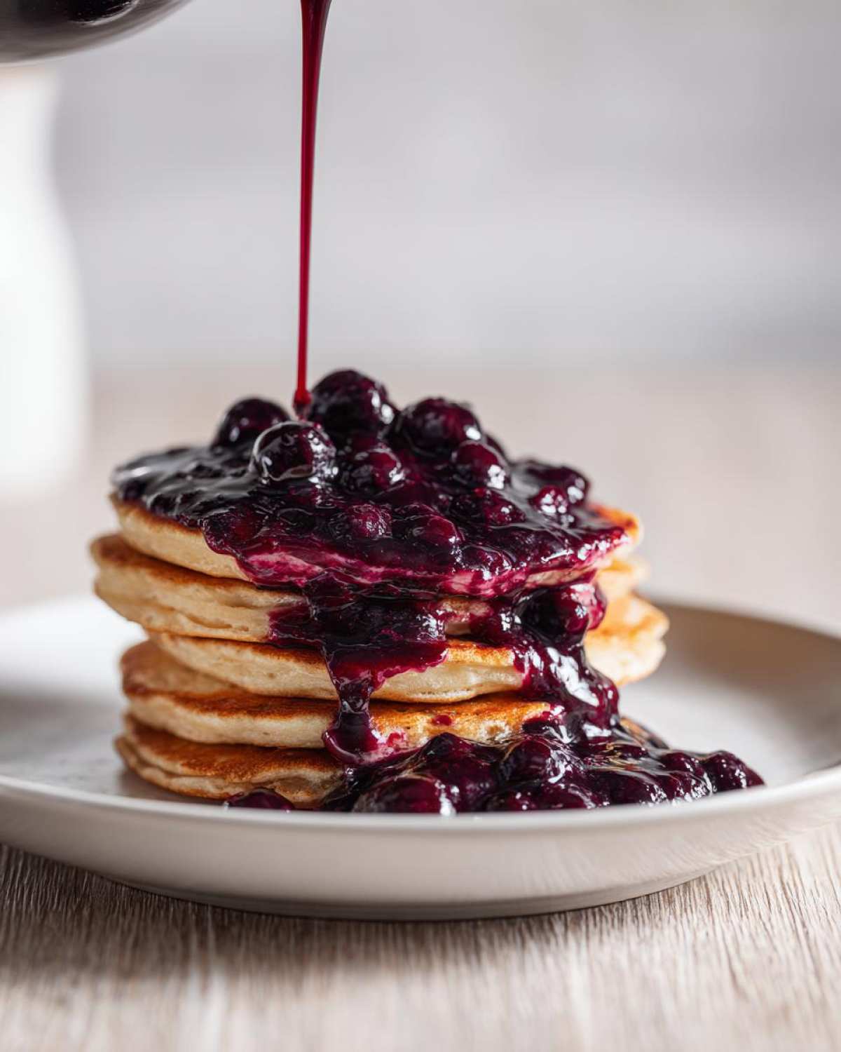 A stream of rich, dark Homemade Blueberry Syrup being poured over a stack of fluffy pancakes.