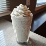 A tall glass of Homemade Vanilla Bean Frappuccino topped high with whipped cream, sitting on a light countertop.