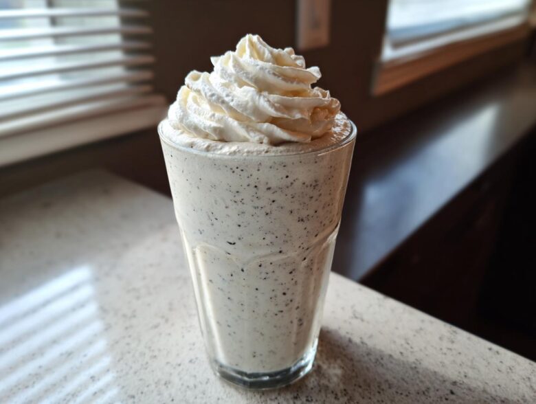A tall glass of Homemade Vanilla Bean Frappuccino topped high with whipped cream, sitting on a light countertop.