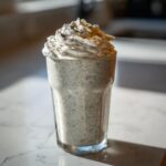 A tall glass filled with a speckled Homemade Vanilla Bean Frappuccino, topped with whipped cream and vanilla bean specks.