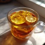 Close-up of a glass mug filled with Honey Lemon Ginger Tea, featuring lemon slices and floating herbs.