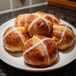 A small pile of freshly baked Hot Cross Buns with Orange Glaze, topped with white icing crosses, sitting on a white plate.
