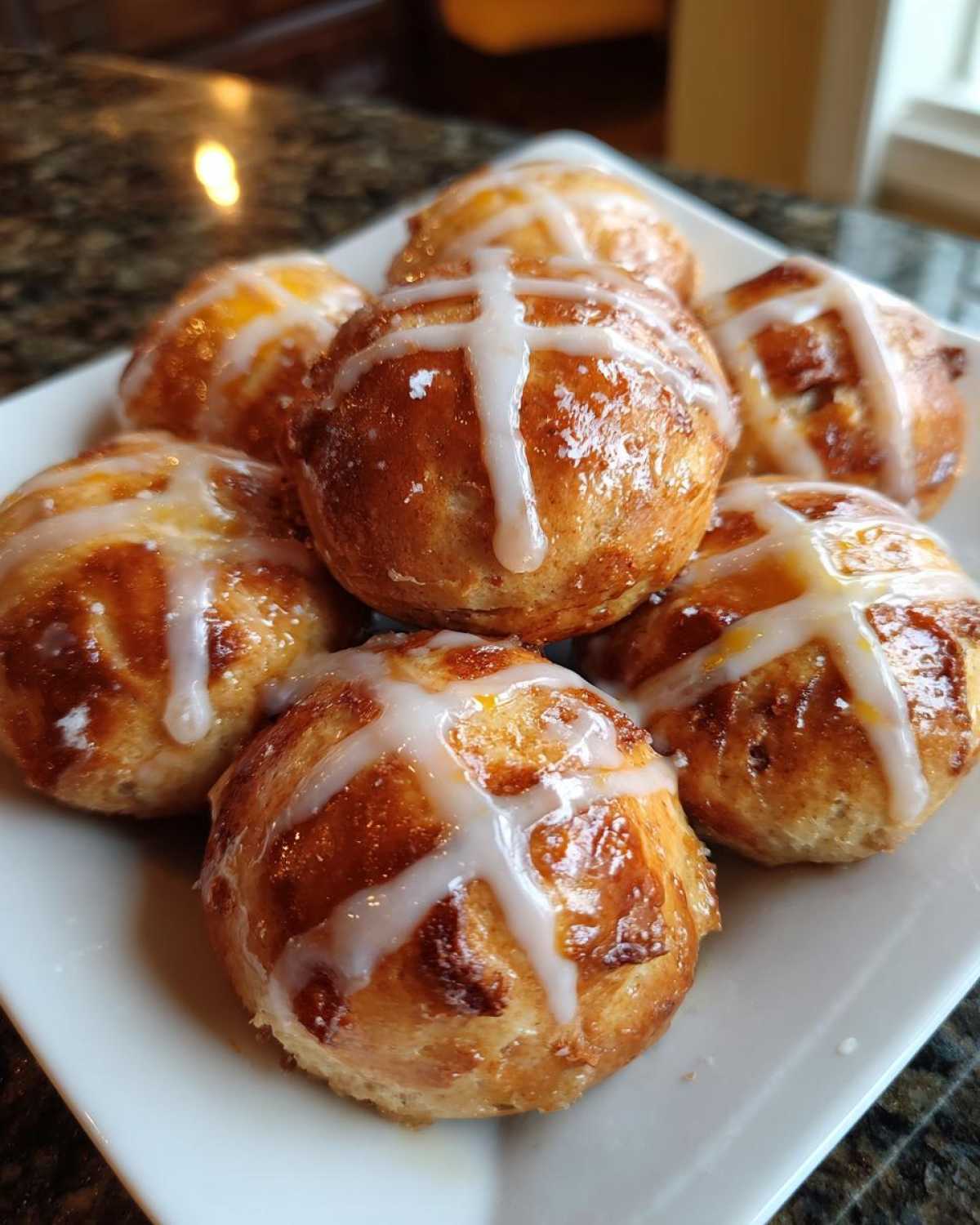 A close-up of several freshly baked Hot Cross Buns with Orange Glaze, drizzled with white icing, served on a white platter.