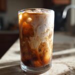 Close-up of an Iced Brown Sugar Oatmilk Latte swirling with coffee and oatmilk over ice in a tall glass.