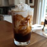 A tall glass filled with a layered Iced Mocha, ice cubes, and topped with whipped cream.