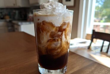A tall glass filled with a layered Iced Mocha, ice cubes, and topped with whipped cream.