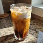 A tall glass of Iced Vanilla Chai Latte with ice cubes and a swirl of cream on top, sitting on a granite counter.
