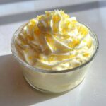 Close-up of a single serving of light yellow Keto Lemon Cheesecake Mousse topped with whipped cream swirls and lemon zest.