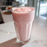 A tall glass filled with a thick, pink Keto Strawberry Cheesecake Smoothie sitting on a white marble counter.