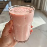 A hand holds a tall glass filled with a thick, pink Keto Strawberry Cheesecake Smoothie near a bright kitchen window.