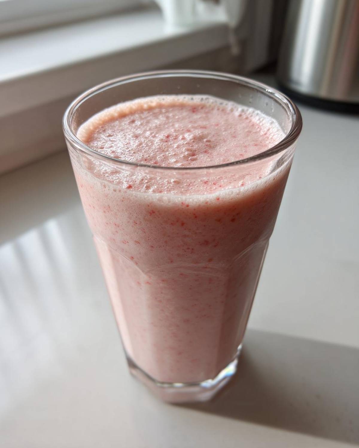 A tall glass filled with a frothy, pink Keto Strawberry Cheesecake Smoothie sitting on a white counter.