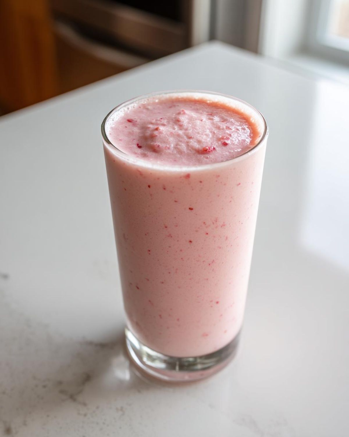 A tall glass filled with a thick, pink Keto Strawberry Cheesecake Smoothie, showing small strawberry flecks.