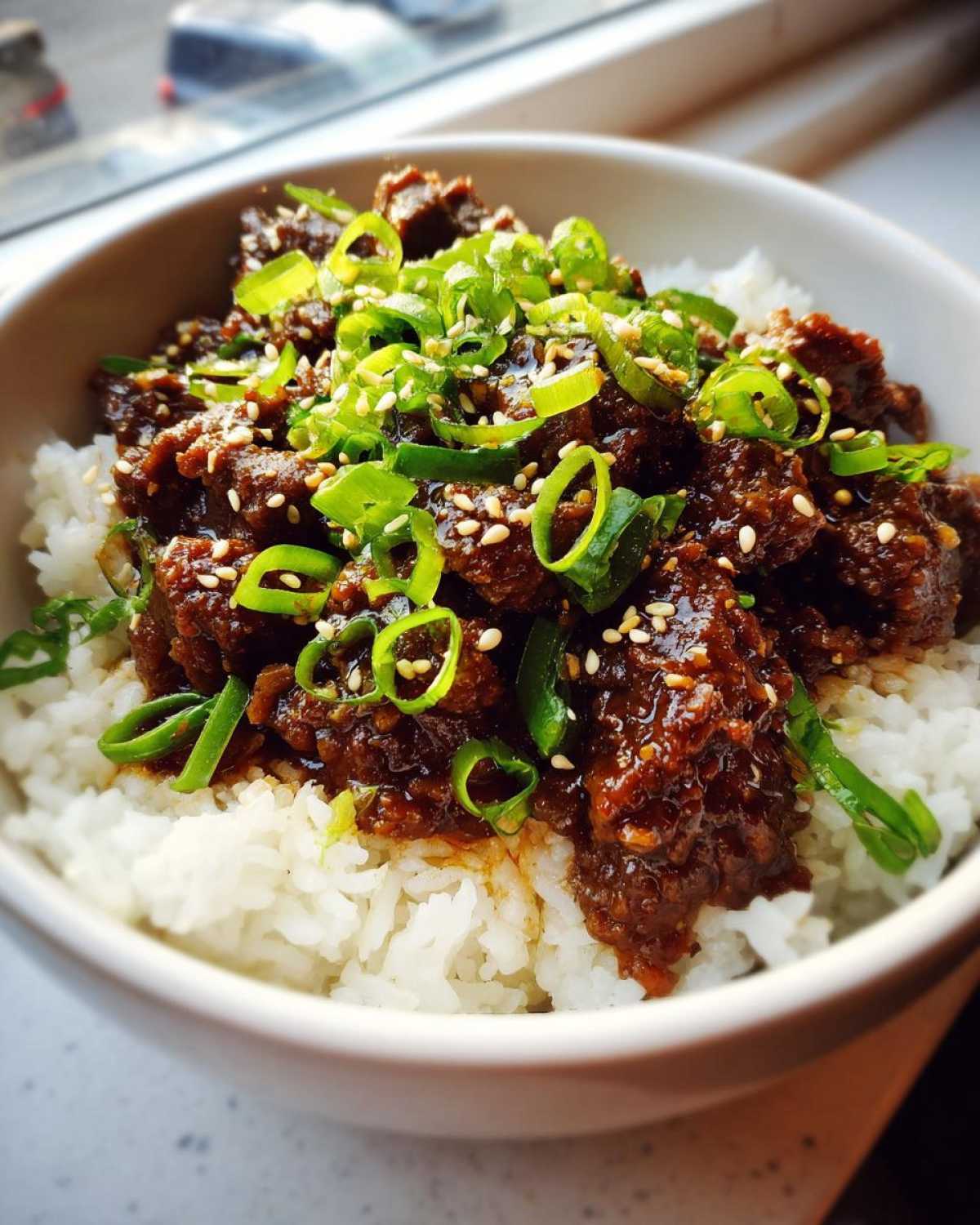 A close-up of Korean Ground Beef Bulgogi Bowls featuring saucy ground beef over white rice, topped with green onions and sesame seeds.