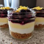 Three individual Lemon Blueberry Cheesecake Jars showing graham cracker crust, cream cheese layers, blueberry topping, and lemon zest.