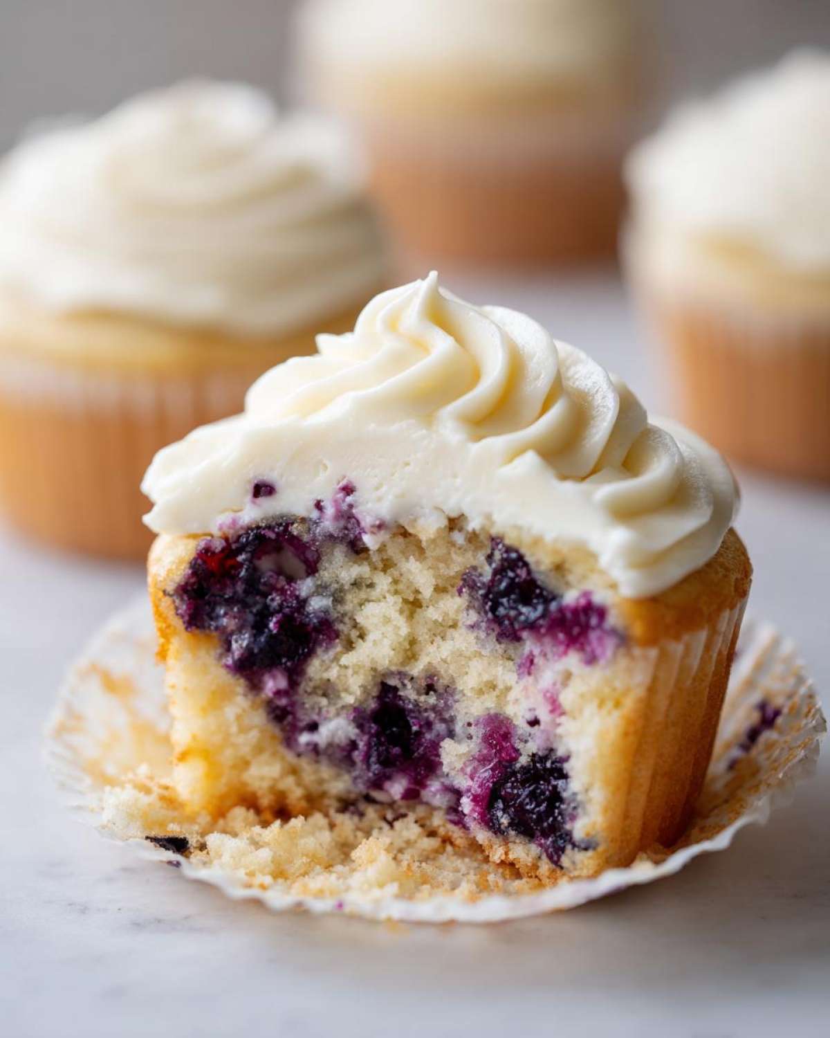 Close-up of a Lemon Blueberry Cupcake cut in half showing moist crumb and blueberries, topped with cream cheese frosting.