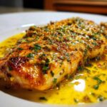Close-up of a perfectly cooked Lemon Garlic Butter Tilapia fillet swimming in rich, yellow butter sauce topped with herbs.