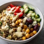 Close-up of a Mediterranean Quinoa Bowl topped with chickpeas, tomatoes, cucumbers, olives, and drizzled with lemon tahini sauce.