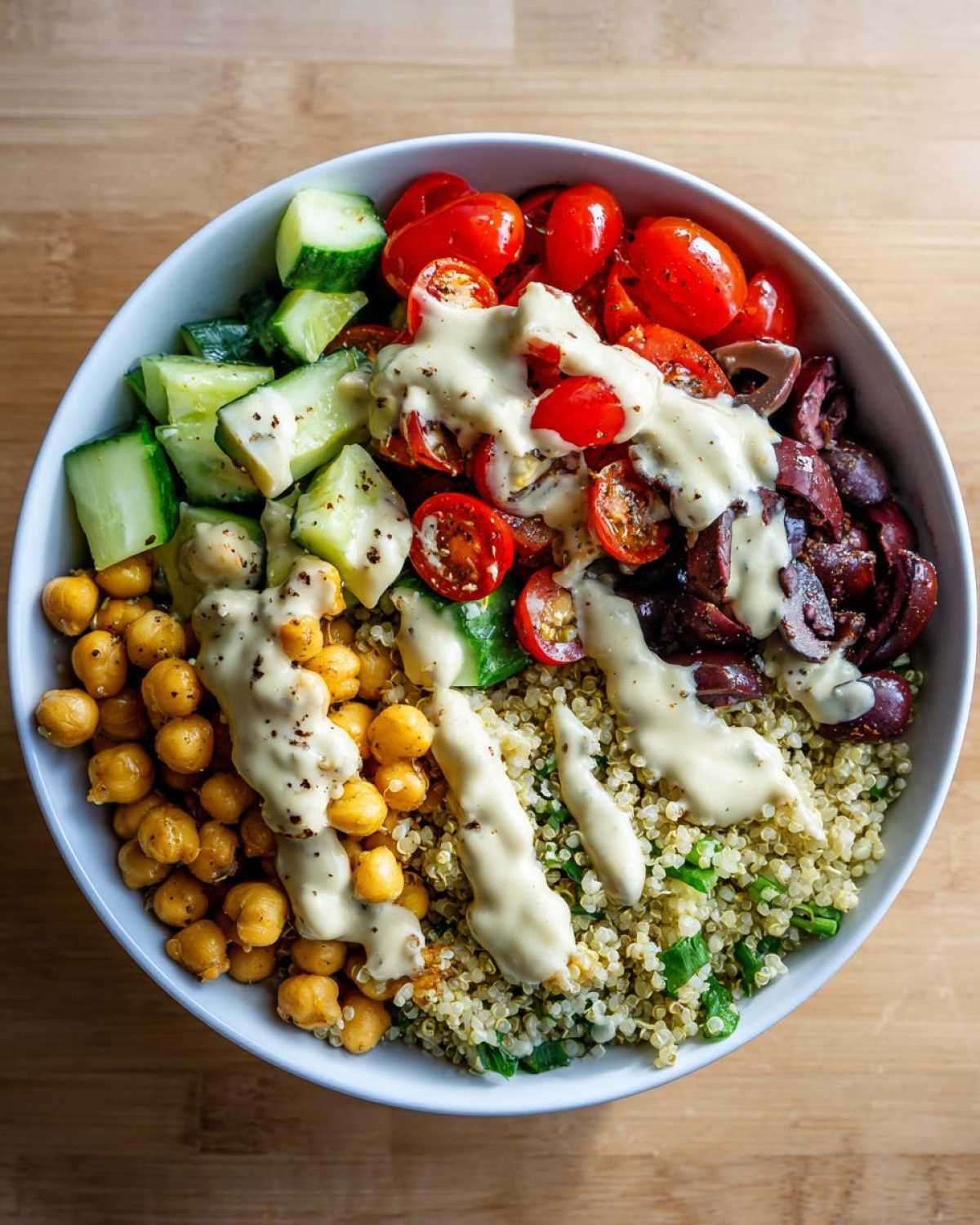 Overhead view of a Mediterranean Quinoa Bowl featuring chickpeas, cucumbers, tomatoes, olives, and drizzled with lemon tahini sauce.