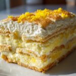 A tall slice of layered No-Bake Lemon Icebox Cake topped with whipped cream and bright yellow lemon zest.