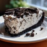 A thick slice of No-Bake Oreo Cheesecake with a creamy filling and dark crust, topped with crushed Oreos.