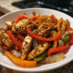 A white bowl filled with seasoned chicken strips, red, yellow, and green bell peppers, and onions from the One-Pan Chicken Fajitas with Peppers and Onions.